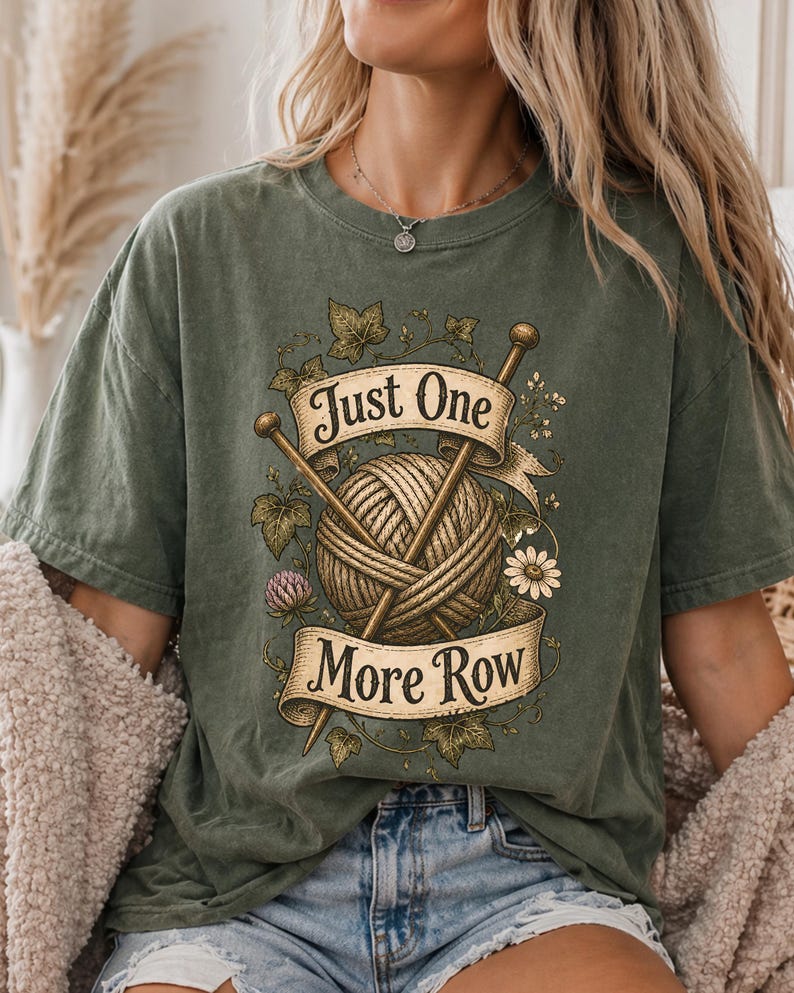 Peut inclure: T-shirt vert olive avec un motif de tricot. Le motif comprend une pelote de laine, des aiguilles &agrave; tricoter et le texte "Just One More Row". Des d&eacute;tails floraux et de feuilles compl&egrave;tent le look inspir&eacute; de l'artisanat.