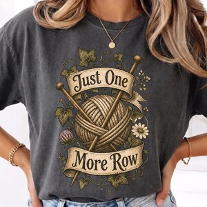 Peut inclure: T-shirt gris fonc&eacute; avec un motif d&eacute;taill&eacute; d'une pelote de laine et d'aiguilles &agrave; tricoter, entour&eacute; d'&eacute;l&eacute;ments floraux. Le motif comprend une banni&egrave;re avec le texte "Just One More Row". Un t-shirt confortable et d&eacute;contract&eacute;.