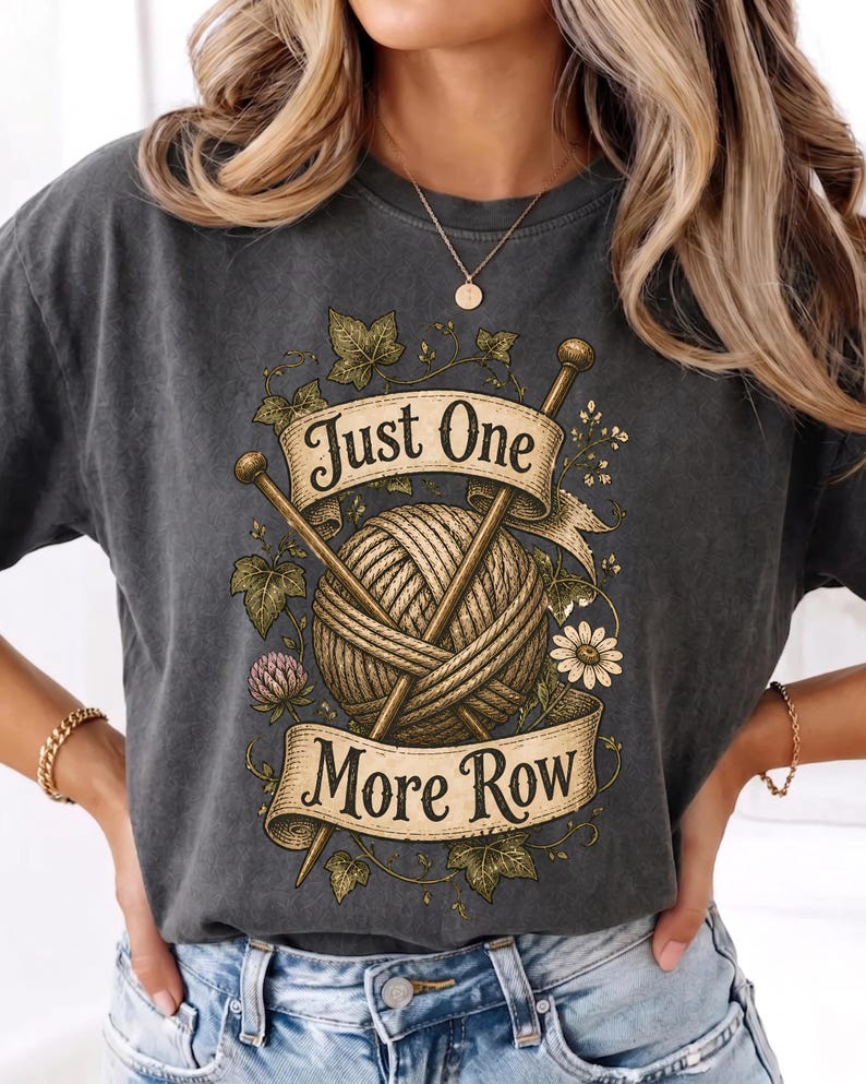 Peut inclure: T-shirt gris fonc&eacute; avec un motif d&eacute;taill&eacute; d'une pelote de laine et d'aiguilles &agrave; tricoter, entour&eacute; d'&eacute;l&eacute;ments floraux. Le motif comprend une banni&egrave;re avec le texte "Just One More Row". Un t-shirt confortable et d&eacute;contract&eacute;.
