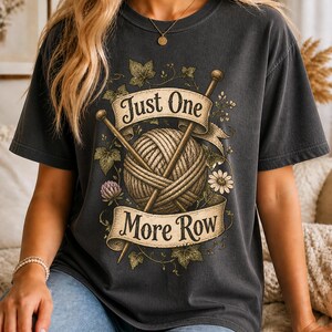 Peut inclure: T-shirt gris fonc&eacute; avec un motif repr&eacute;sentant une pelote de laine, des aiguilles &agrave; tricoter et le texte "Just One More Row". Le motif comprend des &eacute;l&eacute;ments floraux et une banni&egrave;re. Le t-shirt est de style d&eacute;contract&eacute; et confortable.