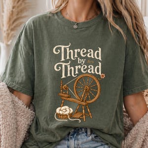 May include: An olive green short-sleeve t-shirt featuring a graphic of a wooden spinning wheel and a basket of yarn. The text "Thread by Thread" is printed in a cream-colored serif font above the illustration, accompanied by a small flower.