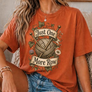 Peut inclure: T-shirt orange br&ucirc;l&eacute; avec un motif d'une pelote de laine, d'aiguilles &agrave; tricoter et le texte "Just One More Row". Le motif est encadr&eacute; d'&eacute;l&eacute;ments floraux. Le t-shirt est de style d&eacute;contract&eacute; et confortable.