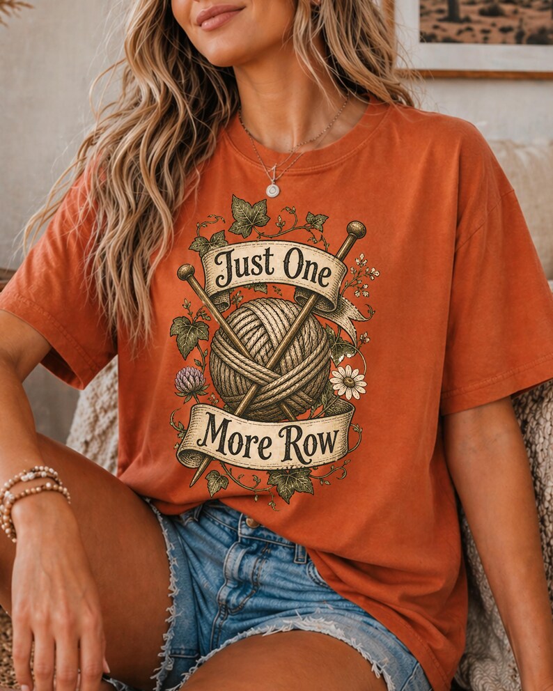 Peut inclure: T-shirt orange br&ucirc;l&eacute; avec un motif d'une pelote de laine, d'aiguilles &agrave; tricoter et le texte "Just One More Row". Le motif est encadr&eacute; d'&eacute;l&eacute;ments floraux. Le t-shirt est de style d&eacute;contract&eacute; et confortable.
