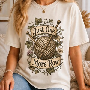 Peut inclure: T-shirt cr&egrave;me avec un motif d'une pelote de laine, d'aiguilles &agrave; tricoter et le texte "Just One More Row". Le motif comprend des accents floraux et de feuilles. Le t-shirt est de style d&eacute;contract&eacute; et confortable.
