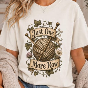 Peut inclure: T-shirt cr&egrave;me avec un motif d'une pelote de laine et d'aiguilles &agrave; tricoter, entour&eacute; de motifs floraux et de feuilles. Le texte "Just One More Row" est affich&eacute; sur une banni&egrave;re. V&ecirc;tement confortable et d&eacute;contract&eacute;.