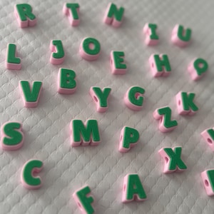 May include: A collection of alphabet beads. Each bead features a green letter with a pink border. The beads are scattered on a white, textured surface. The letters are from the English alphabet.