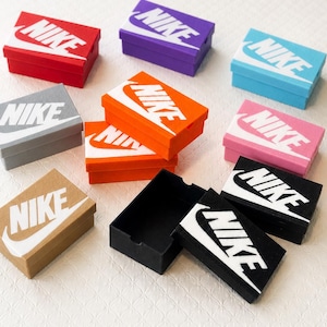 May include: A collection of small, rectangular boxes in various colors, each featuring the Nike logo in white. Colors include red, purple, blue, pink, orange, gray, tan, and black. The boxes are arranged on a white surface.