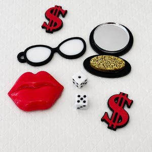 May include: A collection of decorative items including a pair of black-rimmed glasses, two dice, a red lip shape, two red dollar signs, and a compact mirror with a gold-colored surface.
