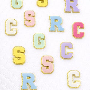Könnte beinhalten: Verschiedene pastellfarbene Buchstabenaufnäher, darunter die Buchstaben G, S, R und C. Jeder Buchstabe hat einen goldglitzernden Rand. Die Buchstaben sind auf einer weißen, strukturierten Oberfläche angeordnet.