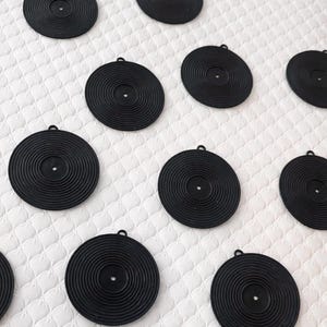 Könnte beinhalten: Eine Ansammlung schwarzer, kreisförmiger Untersetzer mit konzentrischen Rillen, die Miniatur-Schallplatten ähneln. Jeder Untersetzer hat oben eine kleine Schlaufe zum Aufhängen. Die Untersetzer sind auf einer weißen, strukturierten Oberfläche angeordnet.
