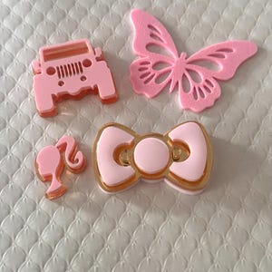 May include: Four pink decorative items: a Jeep, a butterfly, a bow, and a silhouette of a woman's head and hair. The Jeep and bow have gold accents. The butterfly has a detailed wing design. The items are on a white, textured surface.
