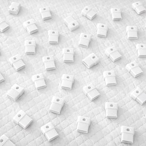 May include: An overhead shot displays numerous small, white, rectangular objects scattered across a textured white surface. Each object has a cylindrical hole on top. The objects are evenly spaced and appear to be made of plastic.