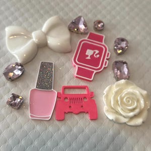 May include: A collection of decorative items: a white bow, pink nail polish, a pink Jeep, a white rose, a pink smartwatch with a silhouette, and pink gemstones. They are arranged on a textured white surface.
