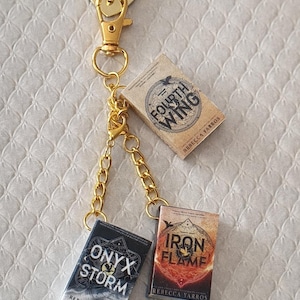 Może przedstawiać: Złoty brelok do kluczy z zawieszką w kształcie serca i trzema miniaturowymi zawieszkami w kształcie książek. Zawieszki książkowe zatytułowane są "Fourth Wing", "Onyx Storm" i "Iron Flame" autorstwa Rebecci Yarros. Brelok jest przymocowany karabińczykiem.
