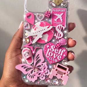 May include: Clear phone case with glitter and pink embellishments. Features a butterfly, airplane, heart, and the words "Self Love Club". Includes a pink Jeep, passport, and dollar sign.