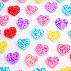 Könnte beinhalten: Ein Muster aus herzförmigen Dekorationen in verschiedenen Farben, darunter Rot, Rosa, Gelb, Blau und Lila. Jedes Herz trägt den Schriftzug "LOVE". Die Herzen sind auf einer weißen, strukturierten Oberfläche angeordnet.