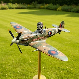 Puede incluir: Maqueta de avión con patrón de camuflaje, con una figura de calavera en la cabina. El avión tiene una hélice y el texto "IRON MAIDEN". Está montado sobre un soporte dorado sobre un fondo de césped verde.