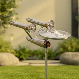 Puede incluir: Una estaca de jardín de metal plateado y cobrizo con la forma de la nave estelar Enterprise de Star Trek. El modelo tiene dos góndolas de curvatura, una sección de platillo y el texto "NCC-1701-D". La estaca está en hierba verde.