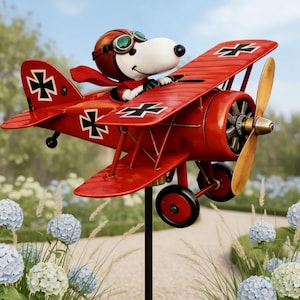 Może przedstawiać: Czerwona dekoracja ogrodowa w kształcie dwupłatowca z postacią psa z kreskówek w kasku i szaliku pilota. Samolot ma czarno-białe oznaczenia i drewniane śmigło. Tło obejmuje kwiaty i zieleń.