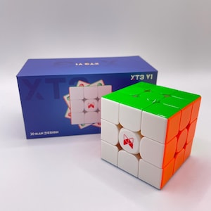May include: A colorful cube puzzle with green, orange, and white sides. The cube has a red logo on the white center squares. A blue box with the text "YT3 V1" and "X-MAN DESIGN" is in the background.