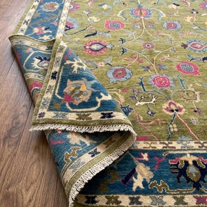 May include: A rectangular area rug featuring a moss green field with a floral and pomegranate pattern in shades of pink, blue, and cream. The rug has a wide blue border with intricate botanical motifs and white fringe along the edges.