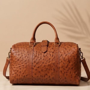 May include: A tan duffel bag with a textured ostrich-skin pattern, featuring two top handles, a central strap with a buckle closure, and side attachments for a shoulder strap. The bag is constructed with brass-toned hardware and stitching.