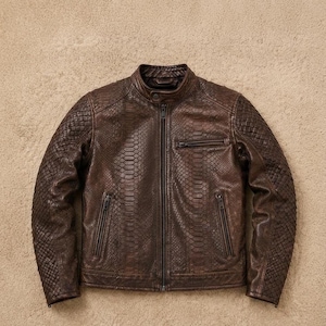 Peut inclure: Une veste en cuir marron foncé présentant un motif texturé en peau de serpent sur les panneaux avant et arrière. La veste comprend un col montant avec un bouton-pression, une fermeture éclair centrale, deux poches latérales zippées et une poche poitrine.