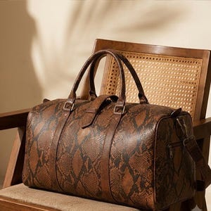May include: A brown and black duffel bag featuring a snakeskin pattern. The bag has two top handles, a central strap with a buckle closure, and a side zipper pocket with metal hardware. The design includes a rectangular shape with rounded edges.