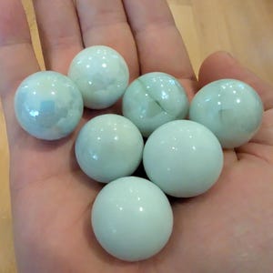 Peut inclure: Sept billes de verre opalescentes de différentes nuances de vert et de blanc sont tenues dans une main ouverte. Les billes ont une finition brillante et sont de forme sphérique. Les billes sont un jouet classique.