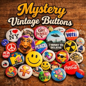 Peut inclure: Une collection de boutons vintage avec divers motifs et textes, dont "I Love NY", "Girl Power", un signe de paix et "I Want to Believe". Les boutons sont disposés sur une surface en bois, avec l'inscription "Mystery Vintage Buttons" en haut.