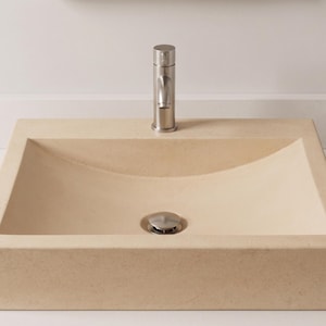 Può includere: Un lavandino rettangolare beige con interno curvo. Sono installati un rubinetto e uno scarico in nichel spazzolato. Il lavandino è realizzato in un solido materiale simile alla pietra ed è appoggiato a una parete bianca.