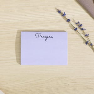 May include: A light purple notepad with the word "Prayers" in cursive script. The notepad is on a light wood surface, with sprigs of lavender and a gold pen nearby. A neutral-toned cloth is also in the image.