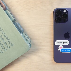 May include: A purple smartphone with a sticker that says "God is good" and "All the time" in a speech bubble. A light green book with tabs labeled with book names is on the left.