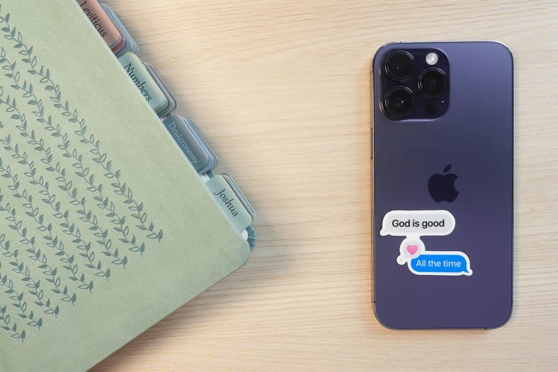 May include: A purple smartphone with a sticker that says "God is good" and "All the time" in a speech bubble. A light green book with tabs labeled with book names is on the left.