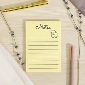 May include: A yellow notepad with the word "Notes" in cursive and a sheep illustration. A gold pen, a pink pen, and sprigs of lavender are arranged around the notepad. A white cloth and a purple cloth are also in the image.