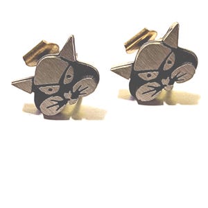 May include: A pair of silver-toned cufflinks featuring a stylized cat face design with black accents. The cat's face has pointed ears and a detailed facial expression. The cufflinks have a gold-colored backing.