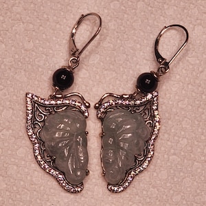 Può includere: Un paio di orecchini pendenti con ali di farfalla intagliate in pietra verde chiaro, incorniciate in metallo color argento con cristalli pavé trasparenti. Ogni orecchino include una perla nera rotonda e una chiusura a monachella.