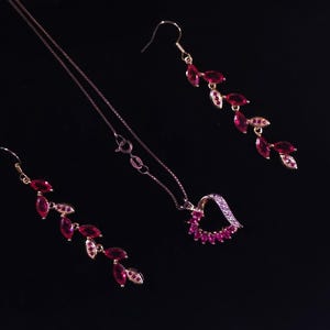 May include: A jewelry set featuring a necklace and earrings. The necklace has a heart-shaped pendant with red gemstones. The earrings and necklace have a gold-colored metal with red and white gemstones. The set is displayed on a black background.