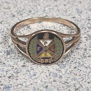 Könnte beinhalten: Ein 10K Goldring mit einem kreisförmigen Emblem auf schwarzem Grund, grünen Lorbeerblättern und einem zentralen Schild, der in vier farbige Bereiche unterteilt ist. Der Schild zeigt die Buchstaben P, A, F, L auf weißen, gelben, blauen und orangen Feldern.