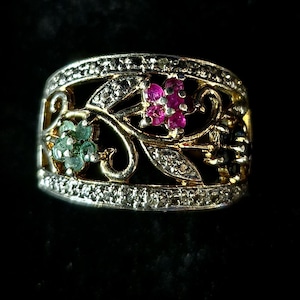 May include: A gold-toned ring with a floral design. The ring features small, round, pink and green gemstones, and is encrusted with small, clear gemstones. The ring is set against a black background.