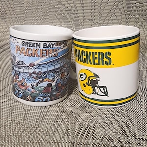 May include: Two white ceramic mugs featuring Green Bay Packers branding. One mug displays a colorful cartoon illustration of a football game with the text GREEN BAY PACKERS. The other mug features the word PACKERS and a yellow football helmet logo.
