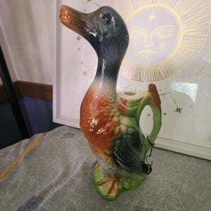 May include: A ceramic duck-shaped pitcher featuring a glossy finish with a dark blue head, a rust-colored chest, and green wings and base. The handle is integrated into the back of the duck, and a small metal ring hangs from the handle area.
