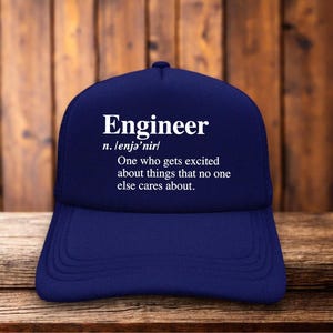 May include: A navy blue baseball cap featuring white printed text that reads: Engineer n. /ɛnʤəˈnɪr/ One who gets excited about things that no one else cares about. The cap has a structured front panel and a curved brim with visible stitching details.