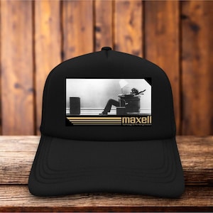 May include: A black baseball cap featuring a rectangular graphic print on the front panel. The print displays the iconic Maxell advertisement image of a person sitting in a chair with the text "maxell" and "Technology for the next generation."