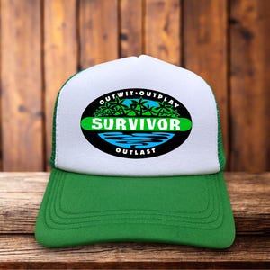 Könnte beinhalten: Eine grün-weiße Trucker-Cap mit einem schwarzen, ovalen Logo auf der Vorderseite. Das Logo zeigt den weißen Schriftzug OUTWIT OUTPLAY OUTLAST sowie das Wort SURVIVOR in Weiß auf einem grünen Balken über einer blauen, stilisierten Wasser-Grafik.