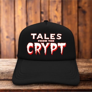 Puede incluir: Una gorra de béisbol negra con el texto TALES FROM THE CRYPT impreso en una fuente blanca y roja que gotea en el panel frontal. La gorra tiene una corona estructurada y una visera curva, manteniendo un color negro sólido en todo el diseño.