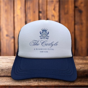 May include: A white and navy blue trucker hat featuring a dark blue crest logo with a crown, the text The Carlyle, A ROSEWOOD HOTEL, and NEW YORK printed in dark blue on the front foam panel, with a solid navy blue brim and mesh back.