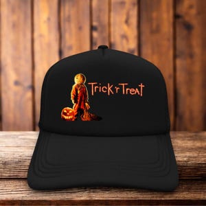May include: A black baseball cap featuring a graphic of the character Sam from the movie Trick 'r Treat standing next to a glowing jack-o'-lantern. The text Trick 'r Treat is printed in an orange, glowing font to the right of the character.