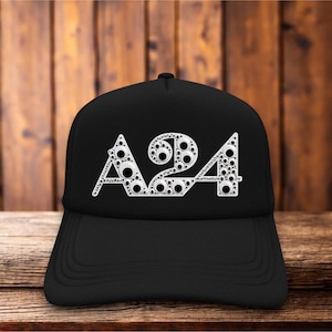 Puede incluir: Una gorra de béisbol negra con el texto A24 en una fuente blanca estilizada. Las letras están rellenas con un patrón de numerosos ojos móviles circulares pequeños, en blanco y negro, de varios tamaños, creando un efecto visual texturizado.