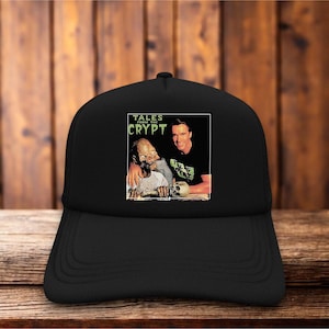 May include: A black baseball cap featuring a rectangular graphic print on the front. The image displays a person with a skeletal figure, with the text "TALES FROM THE CRYPT" printed in green above them. The cap has a structured crown and a curved brim.